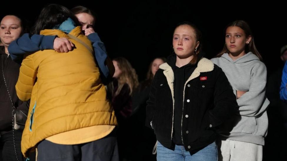 Two people hug each other while three women look on. All of them look sombre  and are wearing light jackets.