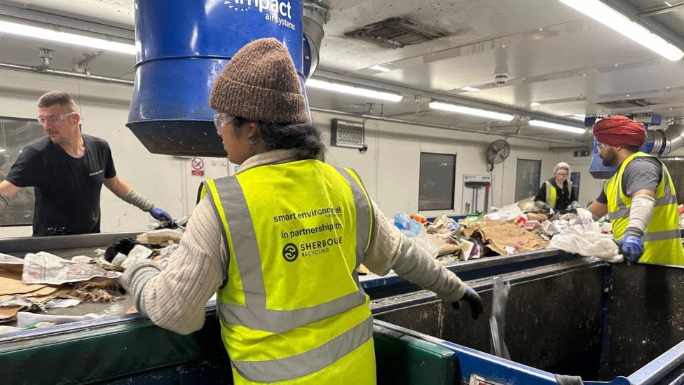 Four workers wearing high-visibility safety vests, gloves and protective eyewear sort mixed waste and recyclables on a conveyor belt inside a recycling facility. Large blue extraction equipment hangs above the sorting line, while strip lights illuminate the industrial workspace.