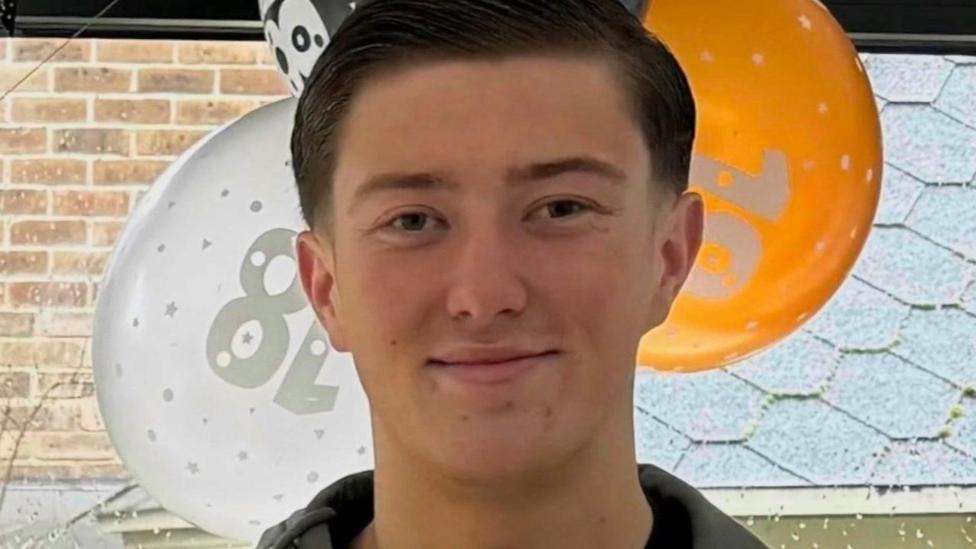 Henry Nowak, 18, standing in front of orange, white and black balloons by a window. He has short dark hair and is smiling and is wearing a blue jacket and a black top underneath.
