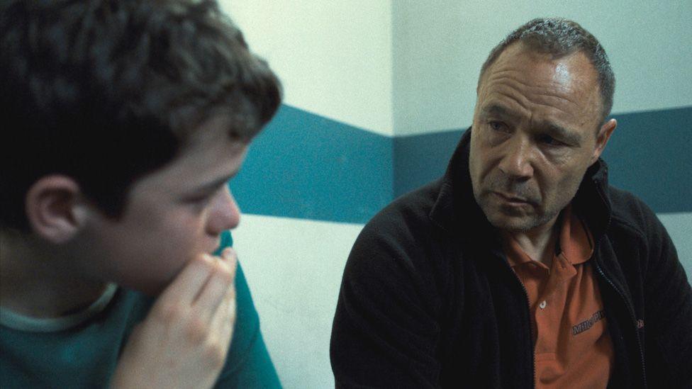 «Adolescence» filmidan olingan kadrda Owen Cooper va Stephen Graham otalik-o‘g‘illik rollarida, politsiya bo‘limida bir-biriga qaragan holda turishibdi. Devorlar oq rangda va bo‘ylab gorizontal ko‘k-yashil (tirol) chiziq o‘tgan.