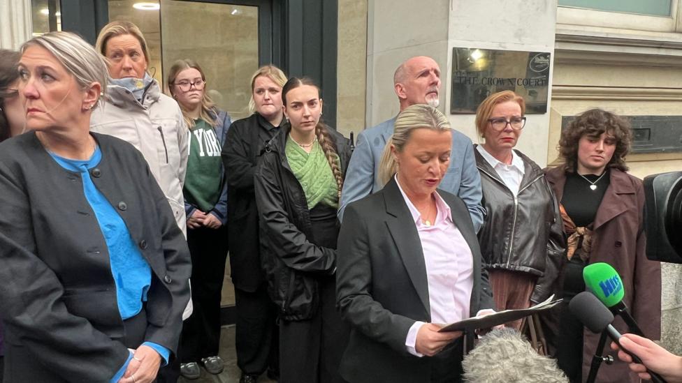 Det Ch Insp Edgeworth to'q rangli kostyum va och pushti ko'ylak kiyib, sud binosi tashqarisida bayonot o'qiyapti, atrofida Jamie Collins'ning oilasi turibdi.