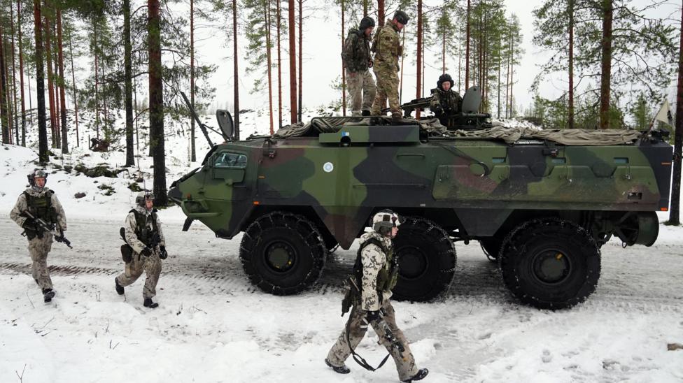 Qorli maydonda uch askar zirhli tank yonida yurmoqda. Uch askar tank ustida o‘tirgan.