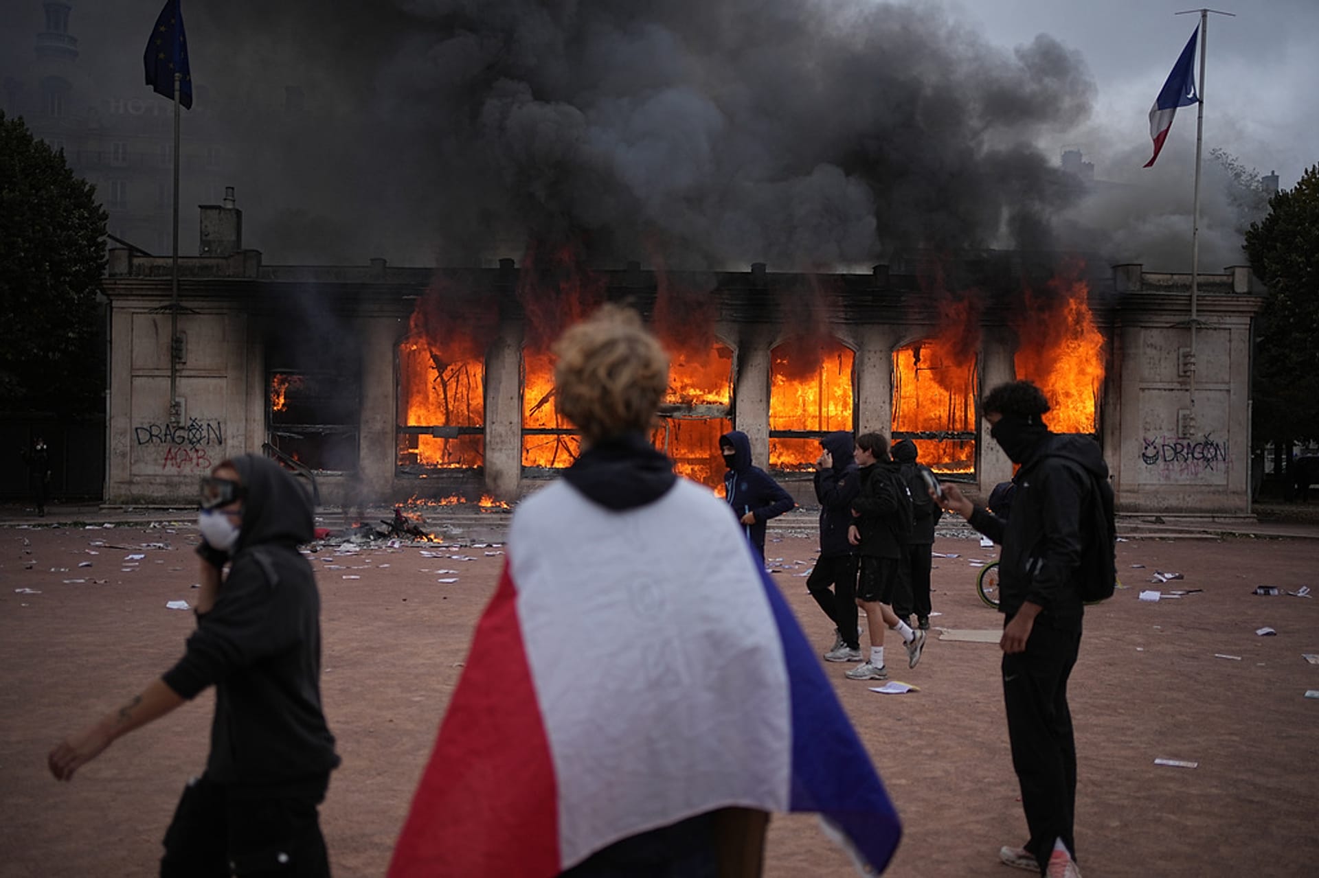 A protester wraps a French flag around them in front of the burning tourism office during a demonstration against rising living costs, Lyon, France, Saturday, Oct. 17, 2026