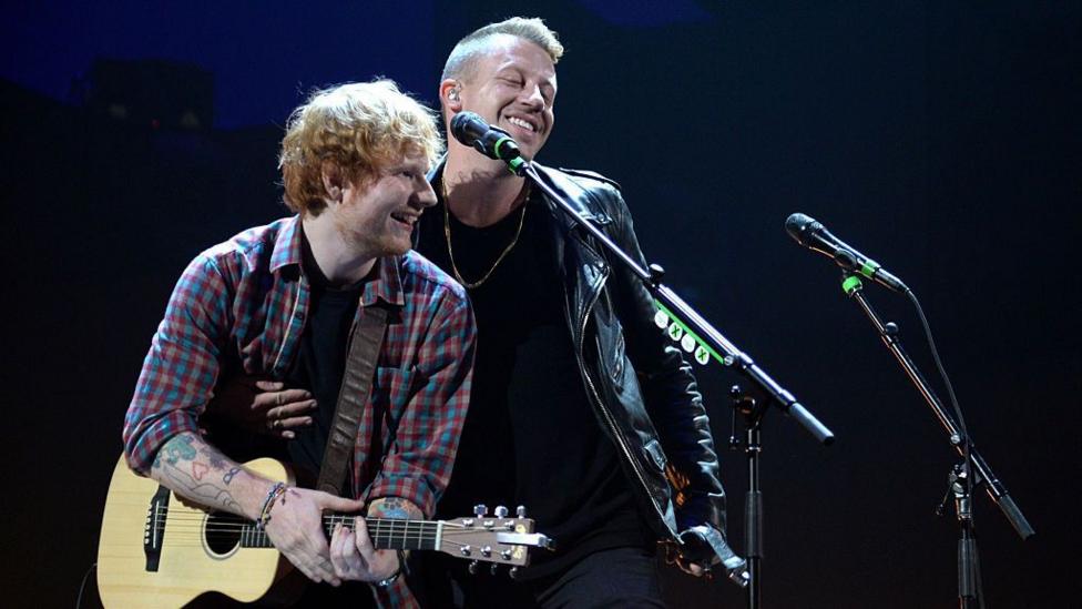 Sheeran holding guitar and smiling over at Macklemore who is also smiling