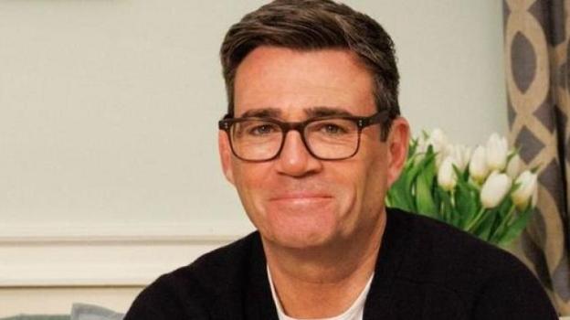 Andy Burnham, wearing glasses with short brown hair, smiles at the camera while wearing a white T-shirt and dark blue cardigan