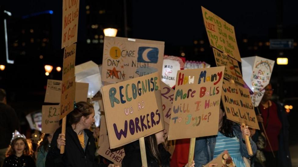 People at a protest at night hold up placards with messages on reading 'recognise us as workers' and 'how many more balls will i have to juggle'.