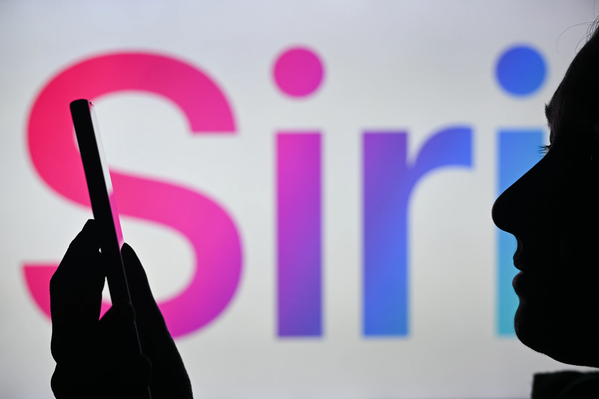 Kompyuter ekranida ko'rsatilgan Siri logotipi oldida telefon ushlab turgan ayol tasviri.