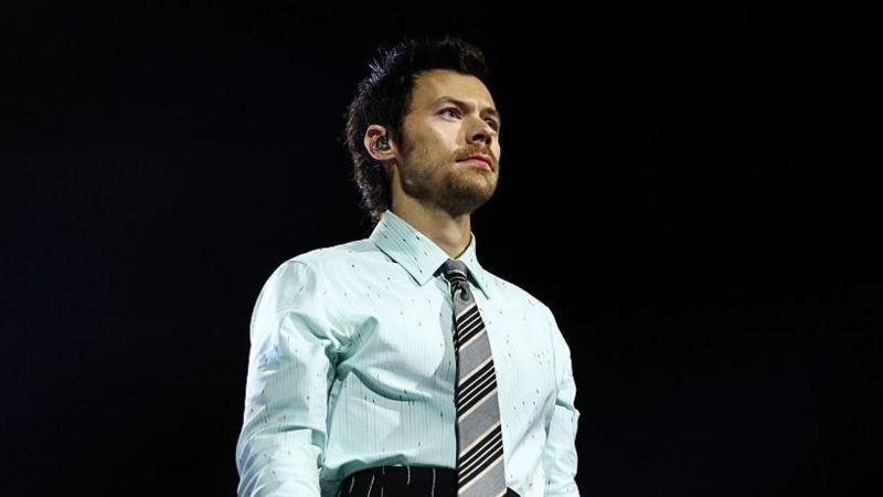 Harry Styles wears a light blue patterned shirt with a grey, white and black striped tie. He has short brown hair and an earpiece and can be seen looking out towards the crowd.