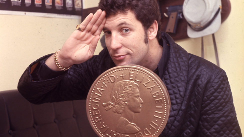 Tom Jones - in the 1960s - giving a salute while he holds an oversized coin