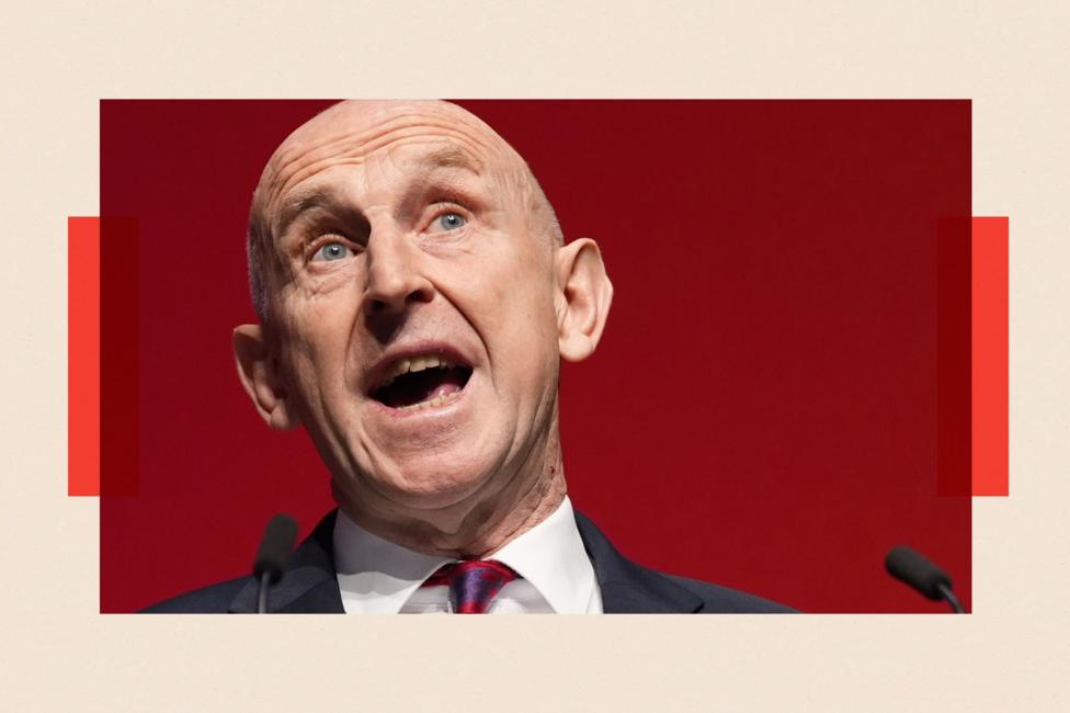John Healey wearing a white shirt with a red and purple tie, speaking at a podium into microphones.