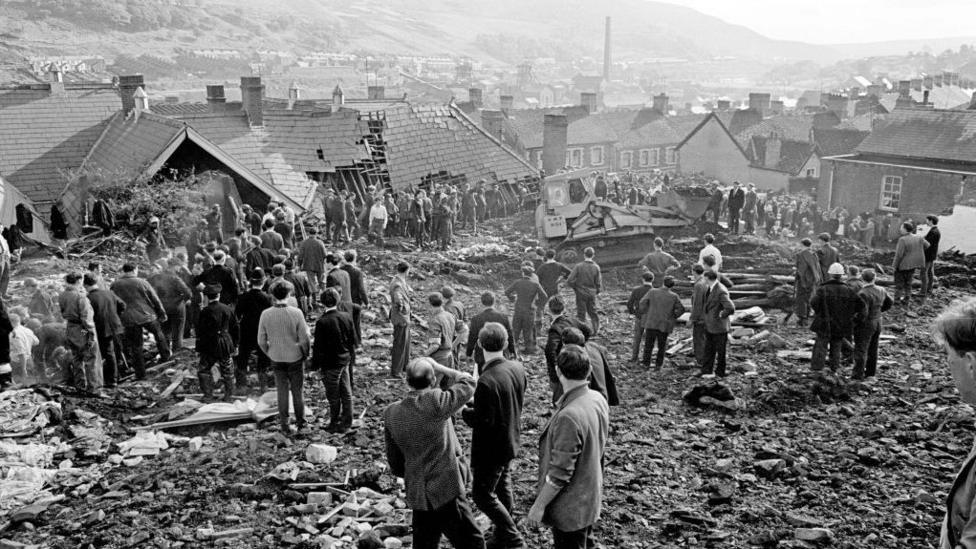 A black and white photo showing rescue workers standing and surveying the damage to the ruined junior school in Aberfan. Coal slurry and debris covers the floor.