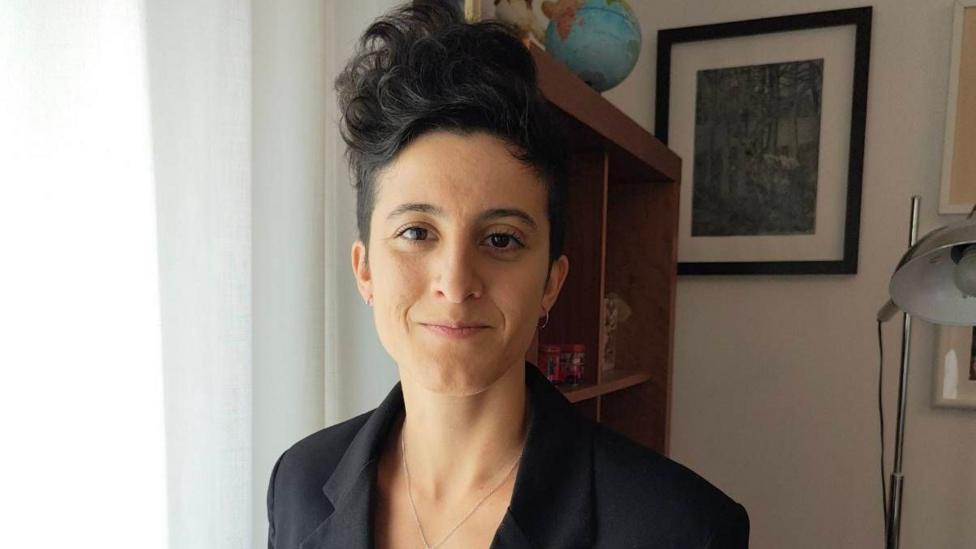 Sofia has short dark hair that is in a quiff style. She is wearing a black blazer and is standing in a room with a lamp and bookshelf in the background.