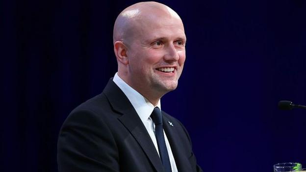 a bald man in a dark jacket, white shirt and dark tie is smiling as he gives a speech