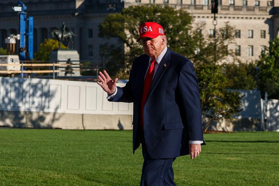 US President Donald Trump walks across a lawn wearing a navy suit, white shirt, red tie and a red cap with USA embroidered on the front. One hand is raised in a wave.