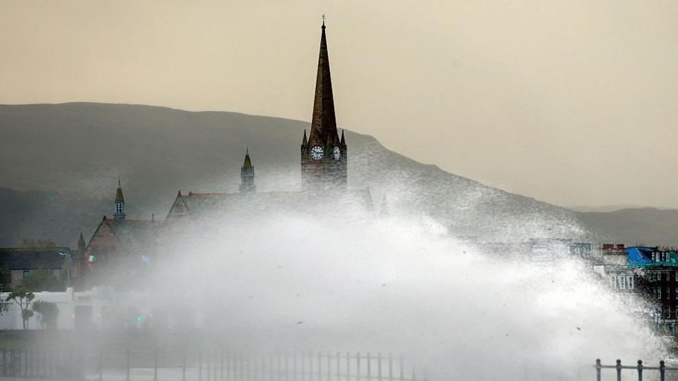 Large waves crash over the promenade in Largs. Buildings, including a clock tower, can be seen behind a huge spray of water, and the sky is overcast.