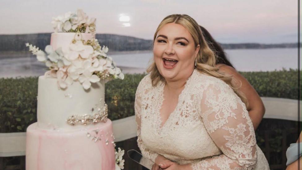 Kayley is wearing a white dress at her wedding and is stood next to a tiered cake, laughing