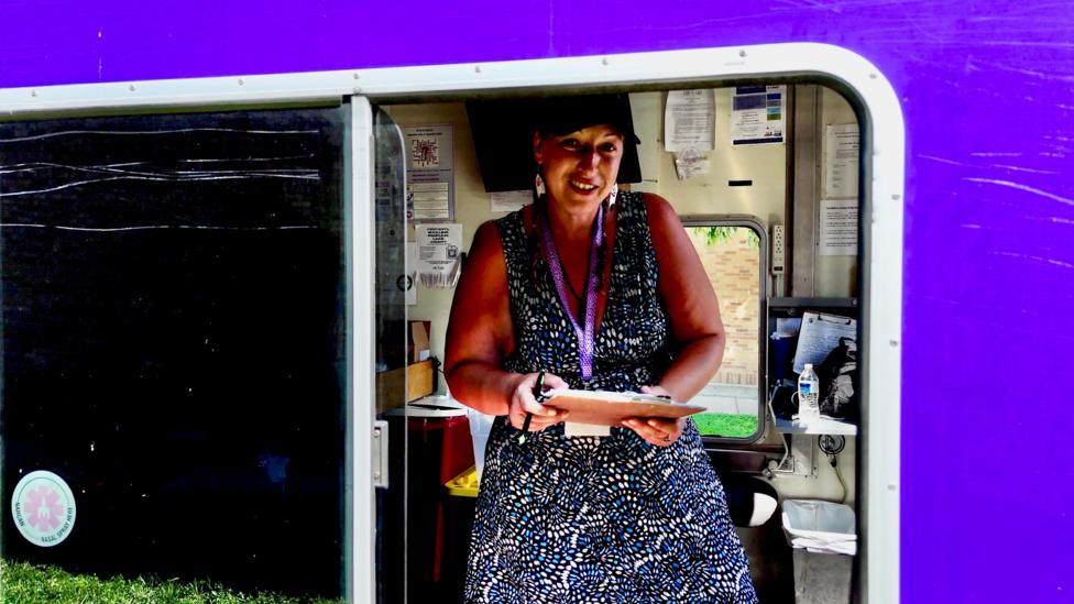 Karen pictured at work. She's wearing a blue-and-white dot dress with a black baseball cap and purple lanyard. She holds a clipboard and smiles at the camera. 