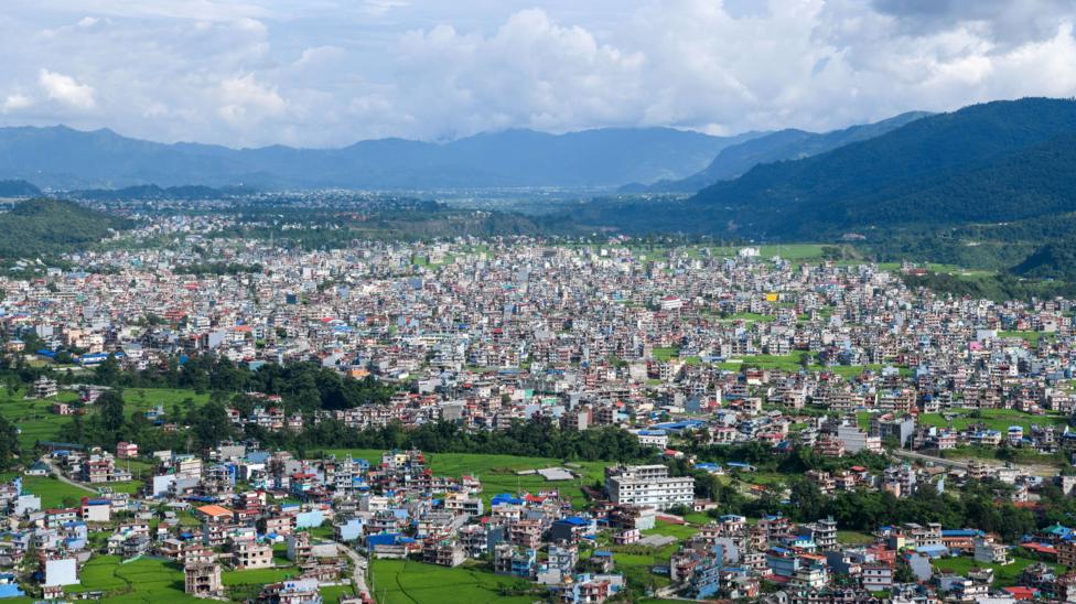 A panoramic view of Pokhara, Nepal, showing a dense spread of homes and low-rise buildings interspersed with bright green fields across a broad valley. Forested hills and mountains surround the city in the distance beneath towering clouds and a hazy blue sky.