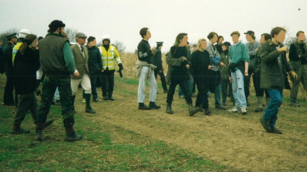 An image showing Andrew Coles and Jessica with their backs to the camera. Both are wearing boots and trousers. They are standing in a farmer's field along with some 20 or so other activists and two police officers wearing motorcycle helmets.