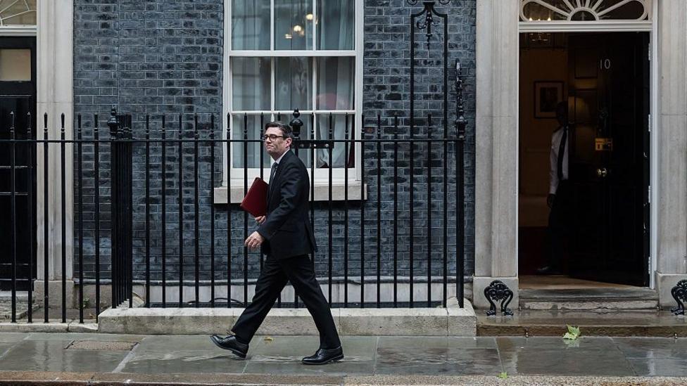 Andy Burnham walks along the pavement in Downing Street in front of the exterior wall and the black Number 10 door.