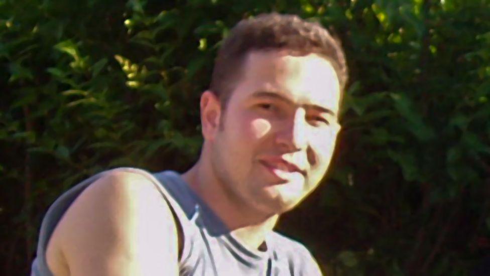 Jean Charles de Menezes smiles at the camera in the sunshine. He has short brown hair and is wearing a pale sleeveless top.
