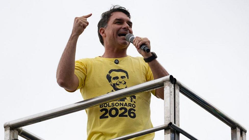 Flavio Bolsonaro otasining yuzi tushirilgan sariq futbolka kiyib nutq so‘zlaydi.