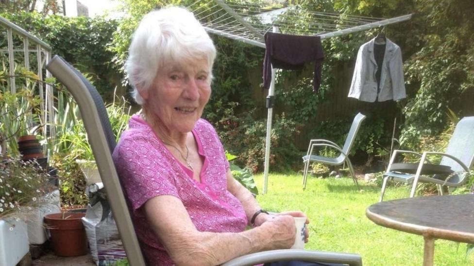 Sheila is sitting in a chair in the garden. She has short, grey hair and is wearing a pink blouse.