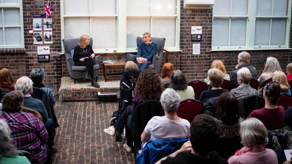 Two people sat on a raised stage leading a talk at a previous Good Grief Weston event. There is an audience of people sat facing them.