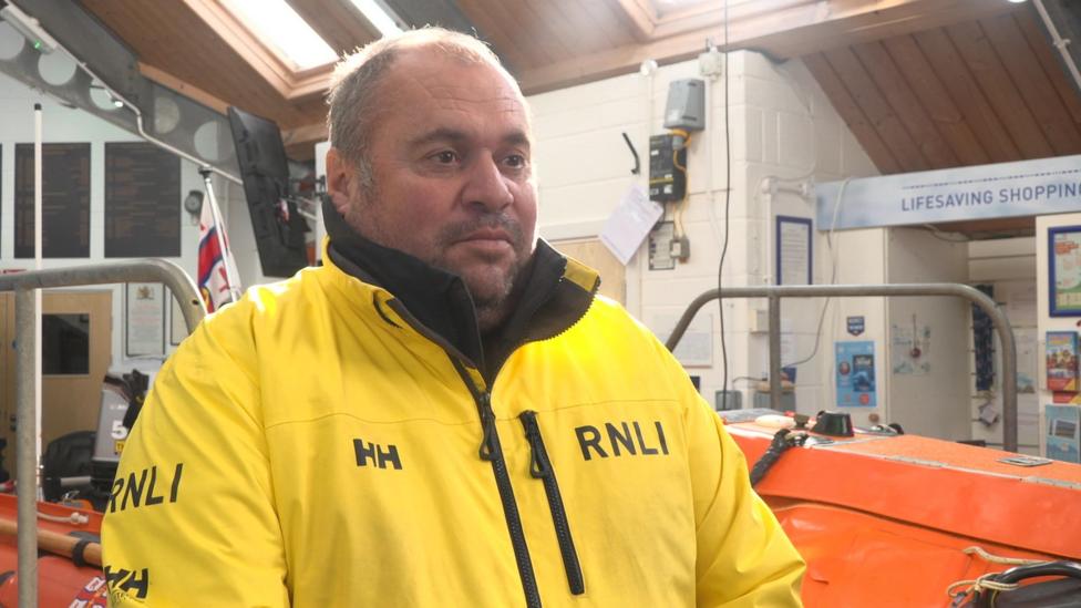 Шкипер RNLI Саймон Маккарти в жёлтой куртке RNLI внутри станции спасательных лодок.