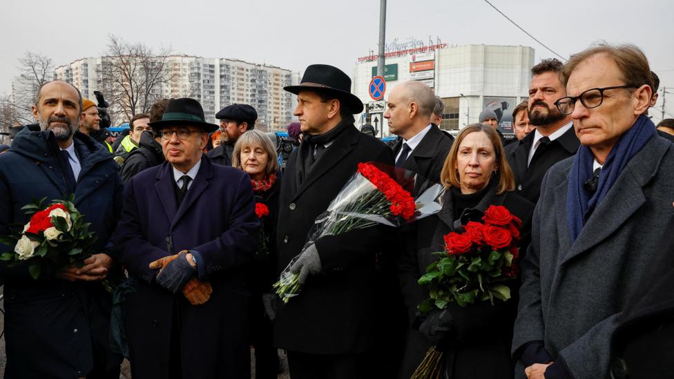 Qora palto kiygan va qizil gul koʻtargan bir qancha odamlar yigʻilgan