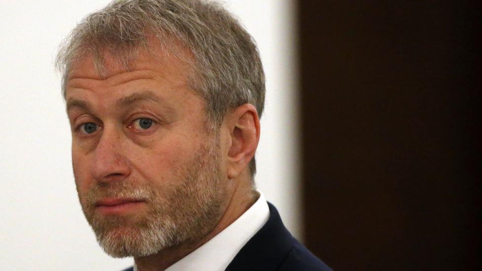 Close-up portrait of Roman Abramovich with short grey hair and a neatly trimmed grey beard, wearing a dark suit jacket and white shirt. He is looking slightly over one shoulder toward the camera. The background is softly blurred, with a light-coloured wall and a dark vertical panel visible.