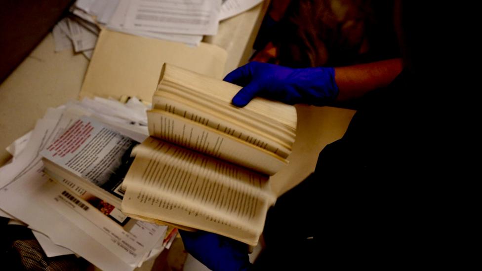 A prison officer flicks through books piled up on a desk 