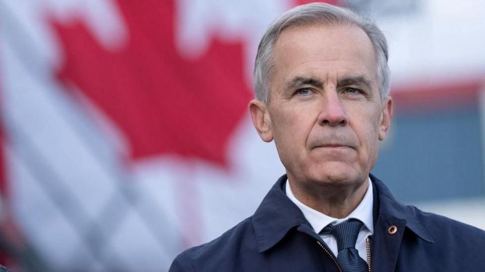Mark Carney, in a dark overcoat and tie, looks straight ahead. Behind him and out of focus, there is a large red and white Canadian flag.
