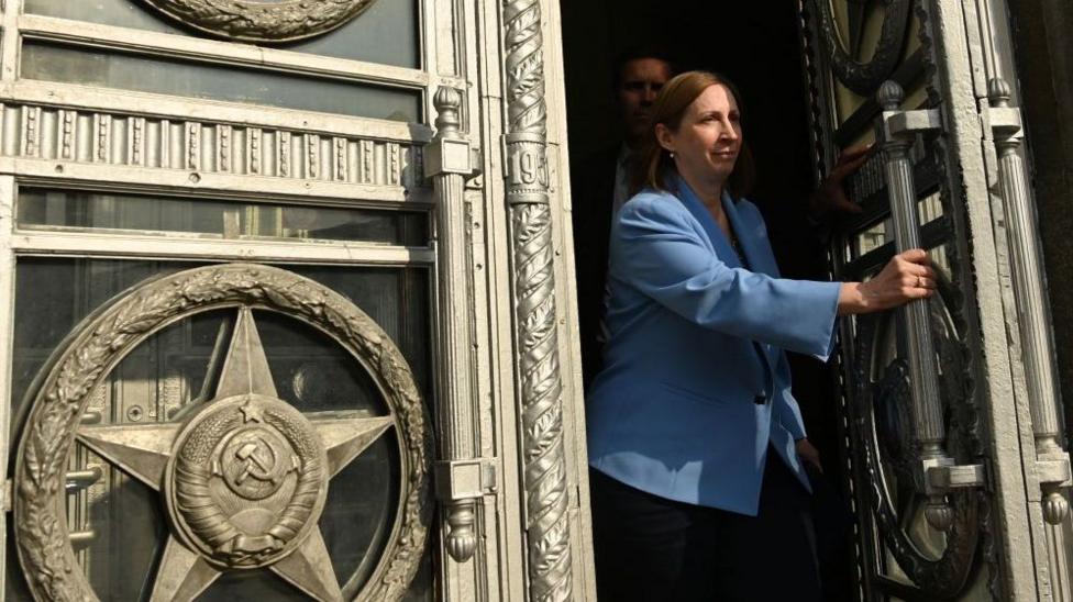 Lynne Tracy katta, nafis qoʻsh eshiklar orasidan yurib kelmoqda. Uning qisqa jigarrang sochlari bor va maviy blazer kiygan. U chap eshikni uzun vertikal dastak yordamida ochmoqda. Eshiklarda, jumladan oʻroq va bolgʻa kabi bronza rangli belgilar oʻyilgan.