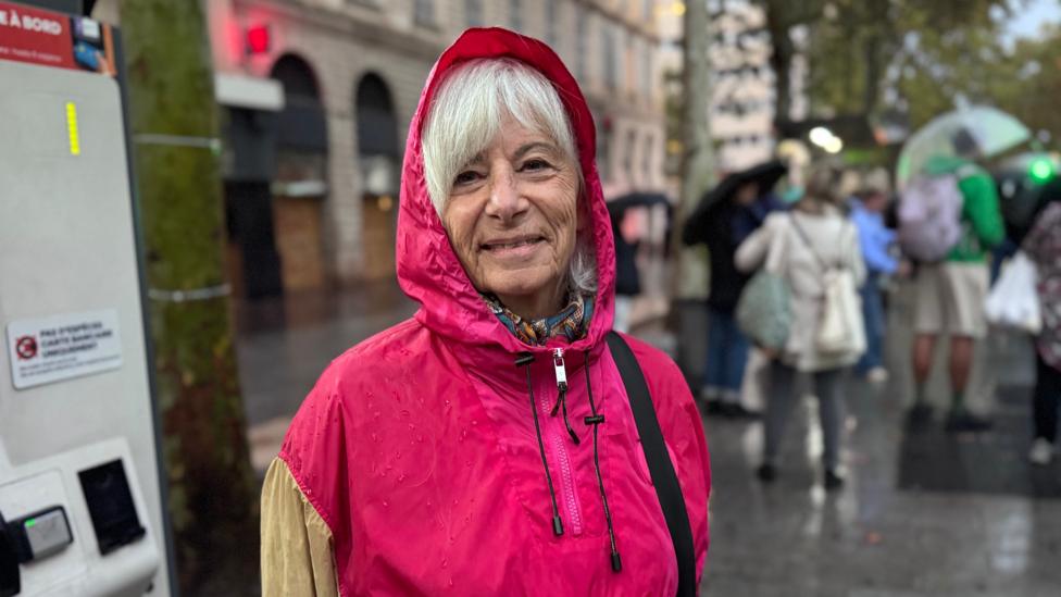 Chantal Bellanger smiles for a photograph in the rain during the daytime. She is an elderly woman with a white fringe, visible underneath the bright pink hood of her raincoat. She is stood at a tram stop, where a number of people are waiting in raincoats and holding umbrellas.