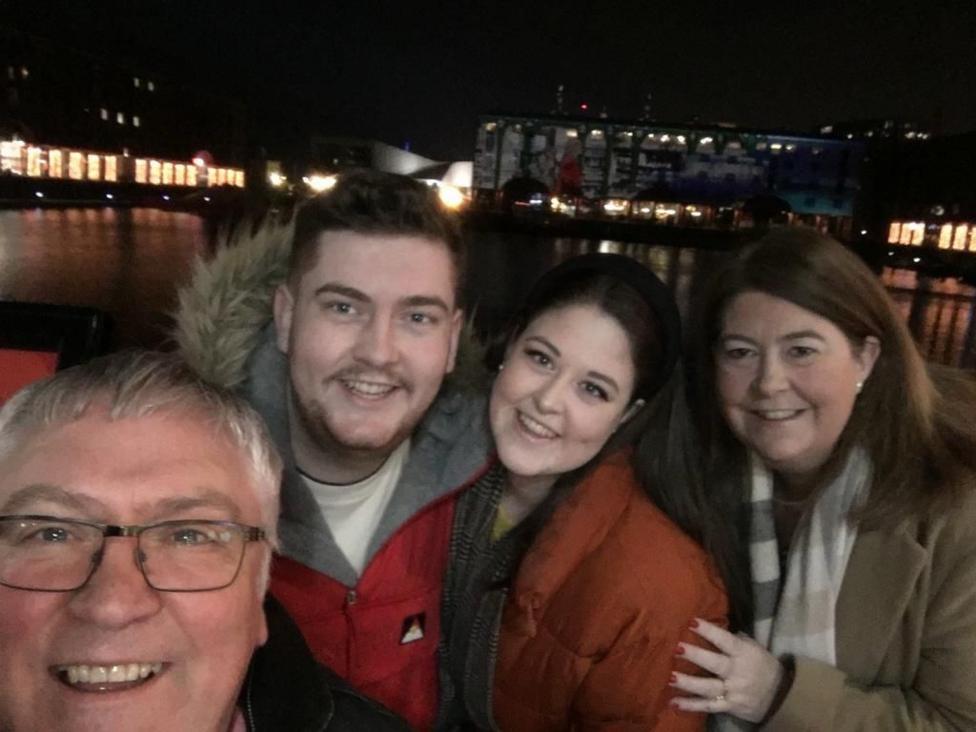 Jon ochiq havoda qorong‘ulikda Jeyms, Emi va Luisa bilan selfi tushgan. Ularning barchasi tabassum qilgan, palto va sharflarda.
