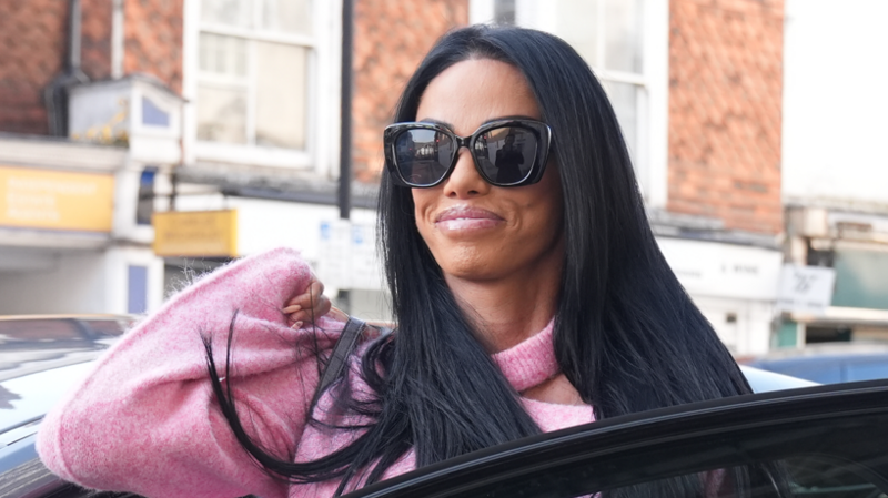 A woman with long black hair, sunglasses and a pink jumper gets out of a car