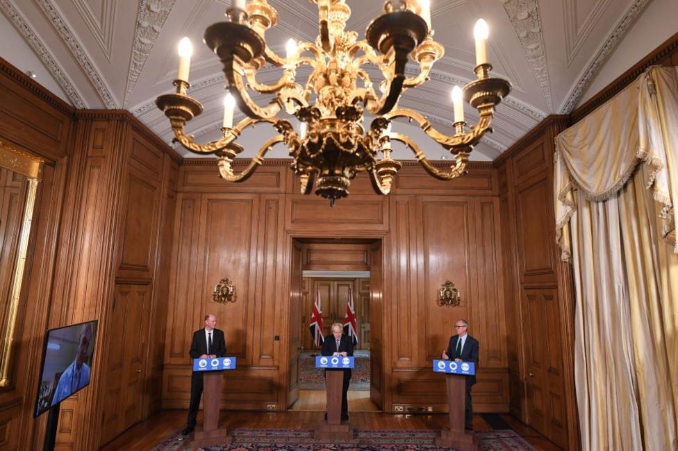 A wood-panelled Downing Street room with cream curtains and a ornate light, with Boris Johnson and his chief medical advisers at podiums during a press conference.
