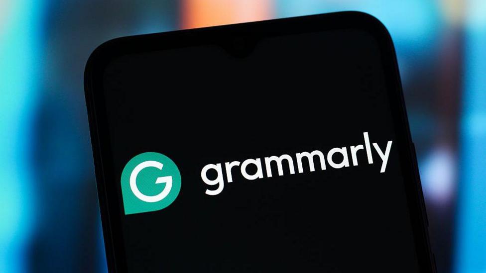 Крупный план экрана смартфона с бело-зелёным логотипом Grammarly на чёрном фоне. Телефон держат на фоне мягко размытого сине-оранжевого боке.