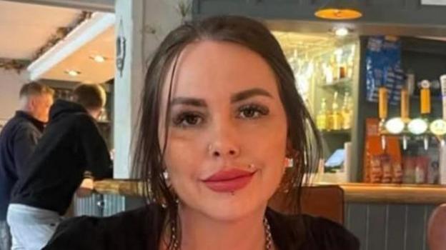 Alisa Webb kameraga qaragan. Uning to'q jigarrang sochlari yuzidan orqaga bog'langan va lab hamda burun piercingi bor