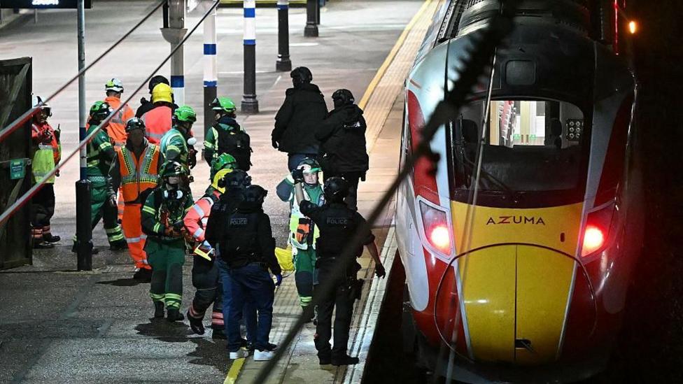 Black-clad police officers and emergency crews wearing green-and-yellow coveralls and helmets on the platform of Huntingdon railway station, where a train has stopped.