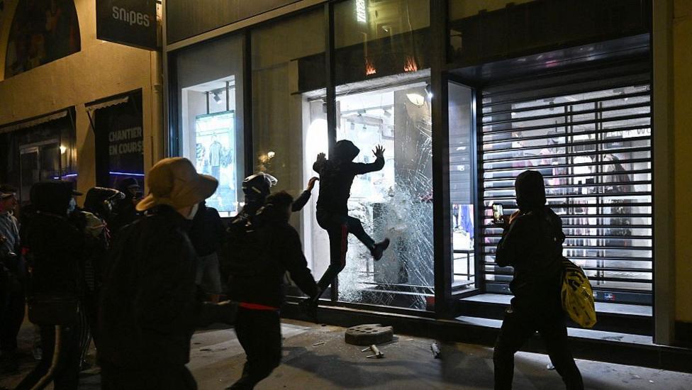 People try to destroy the window of a Snipes store near Chatelet, Les Halles in Paris as riots broke out. There are a group of onlookers looking at someone smashing a shop window. They are all wearing dark clothing and have their faces covered. One person is filming it on a phone.