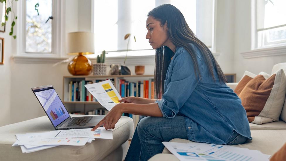 A woman looks at her bills on paper and on a laptop in her sitting room at home