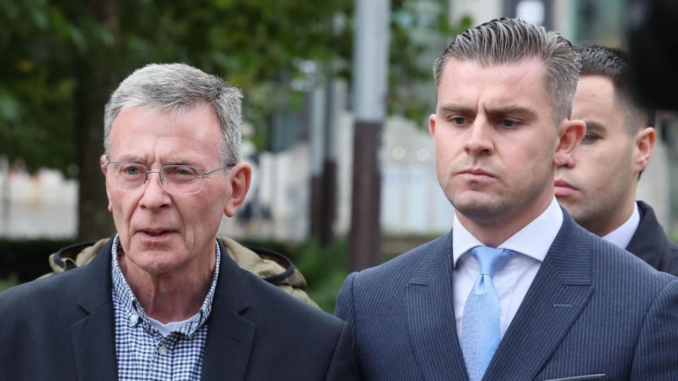 Breandán Mac Cionnaith (chapda) katakli ko‘ylak bilan kostyum kiygan, Darragh Mackin esa to‘q ko‘k kostyum, oq ko‘ylak va och ko‘k galstuk kiygan. Ular sud qarori chiqqanidan so‘ng Oliy sud binosi oldida matbuotga bayonot berayotgan edi.