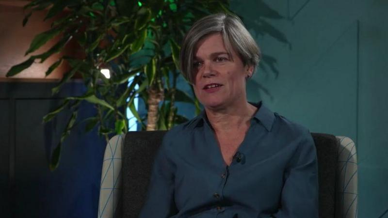 A middle-aged woman with shortish grey hair. She is wearing a turquoise shirt against the backdrop of an indoor plant.