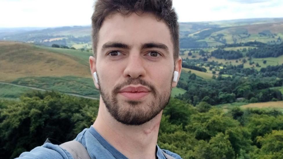 A selfie of William Chapman outdoors, with rolling hills, woodland and patchwork fields stretching across the landscape behind him. He is wearing wireless earbuds and is standing in front of a panoramic countryside view under a bright, cloudy sky.