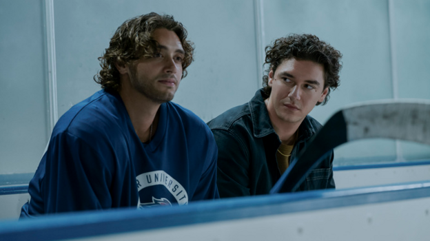 Two men, both with curly brown hair, sat next to each other in the stands of an ice hockey rink. The one on the left is wearing a blue kit, the one on the right looks at him, wearing a black denim jacket.