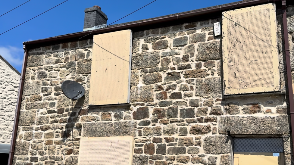 A stone house with the windows and doors boarded up with metal covers