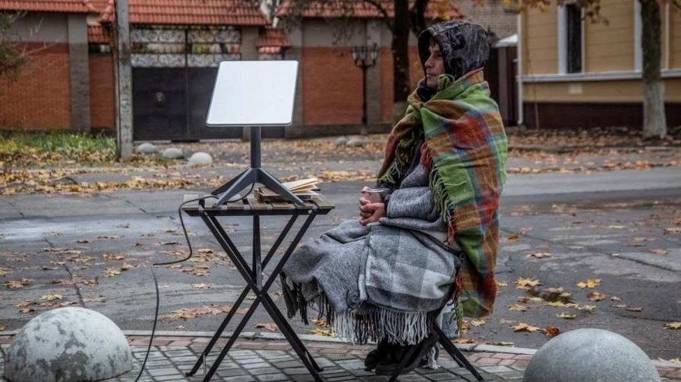 Мужчина, укутанный как минимум в три одеяла и с напитком в руке, сидит на стуле у обочины улицы. Рядом стоит маленький пикниковый столик с терминалом Starlink.