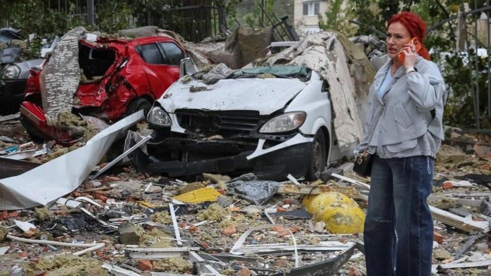 A woman in Ukraine's capital Kyiv talks on the phone near destroyed cars at the site of an apartment building damaged by a Russian strike. Photo: 26 October 2026